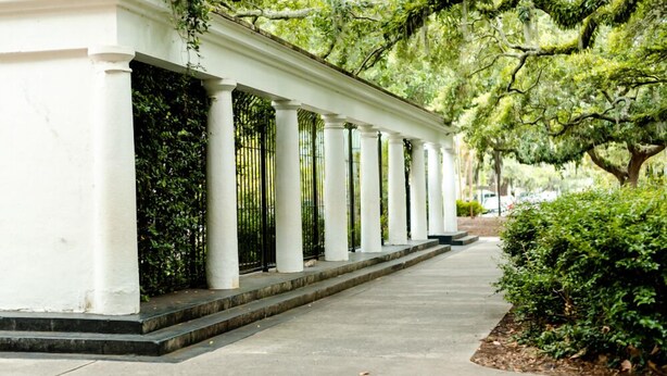 A covered walkway with white columns and a metal railing, surrounded by greenery and trees.