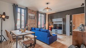 Ein modernes Wohnzimmer mit einem blauen Sofa, einem Esstisch mit Stühlen, einem Fernseher und einer Küchenzeile mit Schränken.