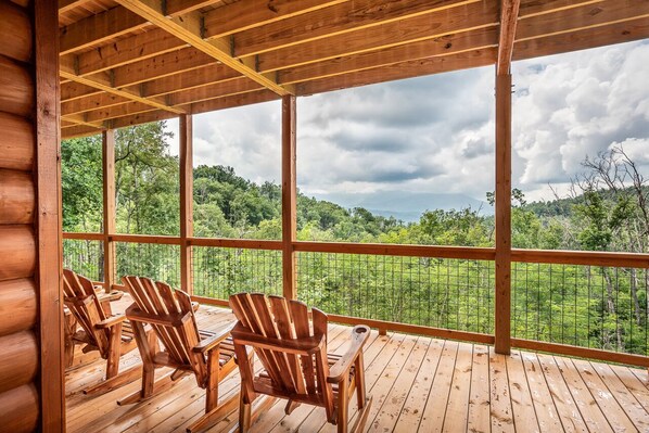 A wooden deck with Adirondack chairs and a large window overlooking a forested landscape. - Four Bears: Amazing View, Private, Arcade, Screened in Porch with Wood Burning (Gatlinburg)