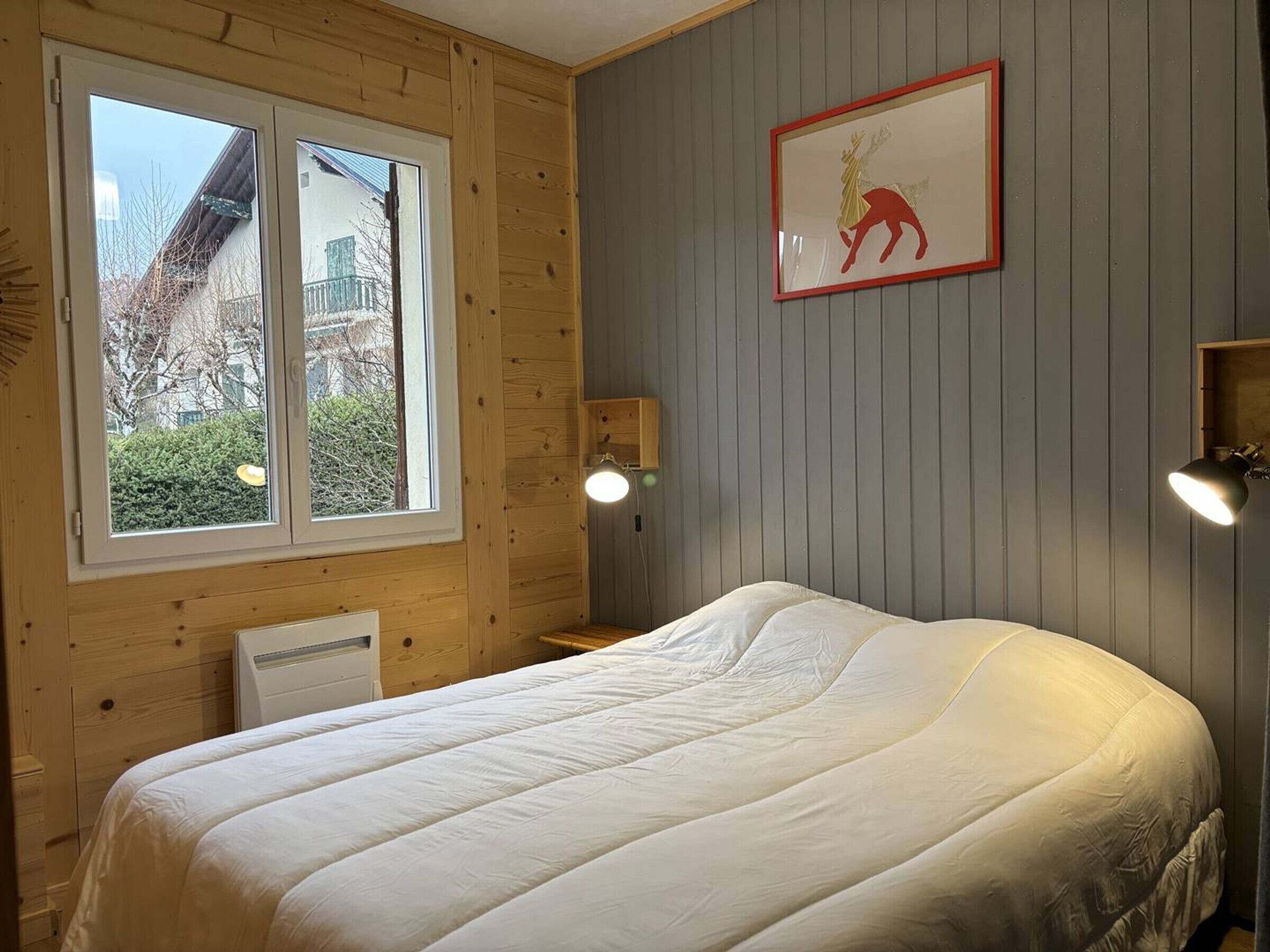 A bedroom with a bed, a window, a wall-mounted lamp, and a framed picture.
