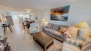 A living room with a sofa, armchairs, a coffee table, a dining table with chairs, a kitchen area, and a painting of a sunset. - Venus 308 overlooking pool Easy Beach Access by RedAwning (Fort Walton Beach)