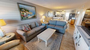 Ein Wohnzimmer mit einer Couch, einem Couchtisch und einem gemusterten Teppich. An der Wand hängt ein Gemälde mit einer Küstenlandschaft.