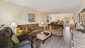 Ein Wohnzimmer mit einer Couch, einem Hocker und Sesseln, ein Essbereich mit Tisch und Stühlen sowie eine Küche im Hintergrund.
