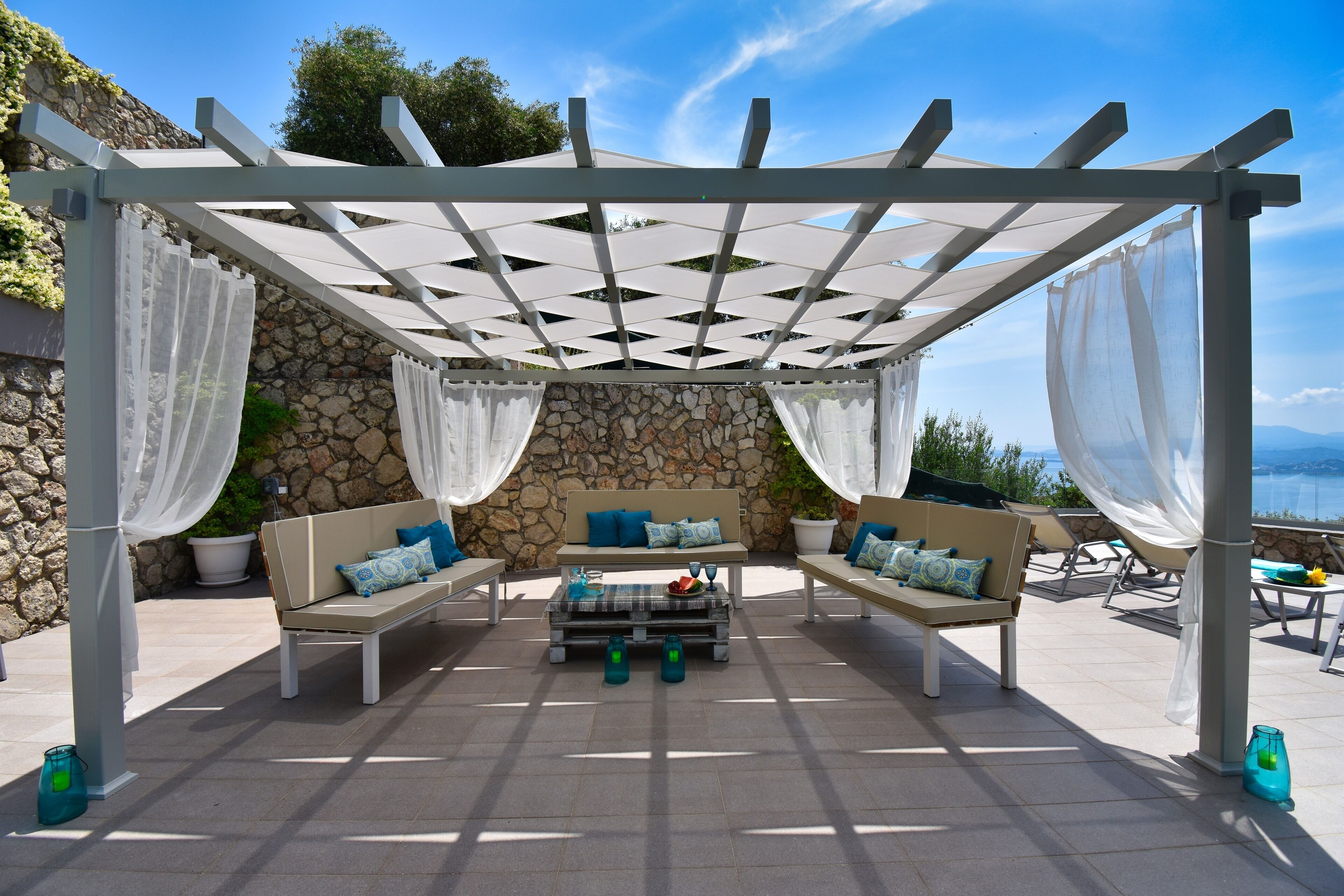 Een zitgedeelte buiten met een stenen muur, een pergola met witte stof en uitzicht op zee.