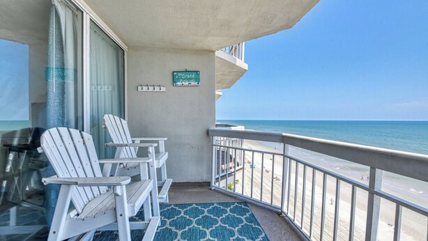 Een balkon met witte Adirondack stoelen, een blauw tapijt met patroon en uitzicht op het strand.