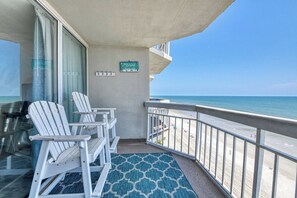 A balcony with white Adirondack chairs, a blue patterned rug, and a view of the beach. - Water's Edge 708 - One Bedroom, Two Bathroom, Oceanfront Condo (Garden City Beach)