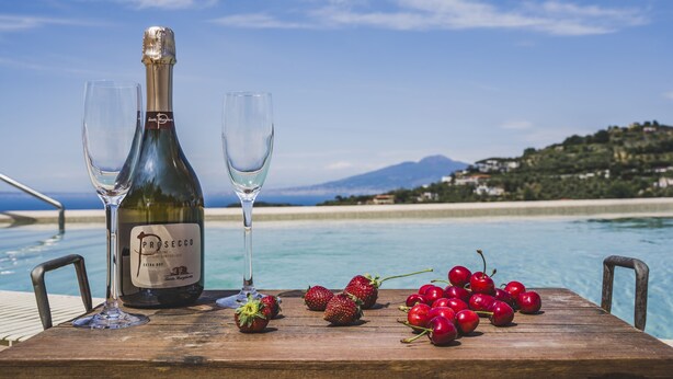 Une table en bois, sur laquelle sont posés une bouteille de Prosecco et deux verres, est entourée de fraises et de cerises. De là, on a une vue imprenable sur une piscine, une plage et des montagnes.