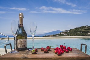 Et træbord med en Prosecco-flaske og to glas, omgivet af jordbær og kirsebær, med udsigt til en pool og et smukt landskab med strand og bjerge.