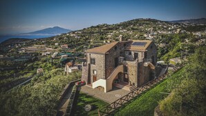 Un bâtiment en pierre, doté d’un toit en tuiles et d’un petit escalier menant à une porte, le tout entouré de verdure et offrant une vue sur une ville située sur une colline.