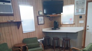 Habitació amb parets revestides de fusta, un televisor de pantalla plana muntat sobre una zona de bar, cadires tapissades de color verd, una taula rodona petita i una finestra amb persianes.