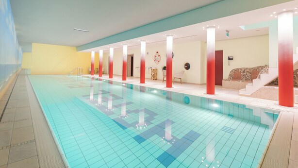 A swimming pool with tiled floor and walls, surrounded by red pillars, and a staircase leading to a seating area. - König Ludwig Designhotel (Altötting)