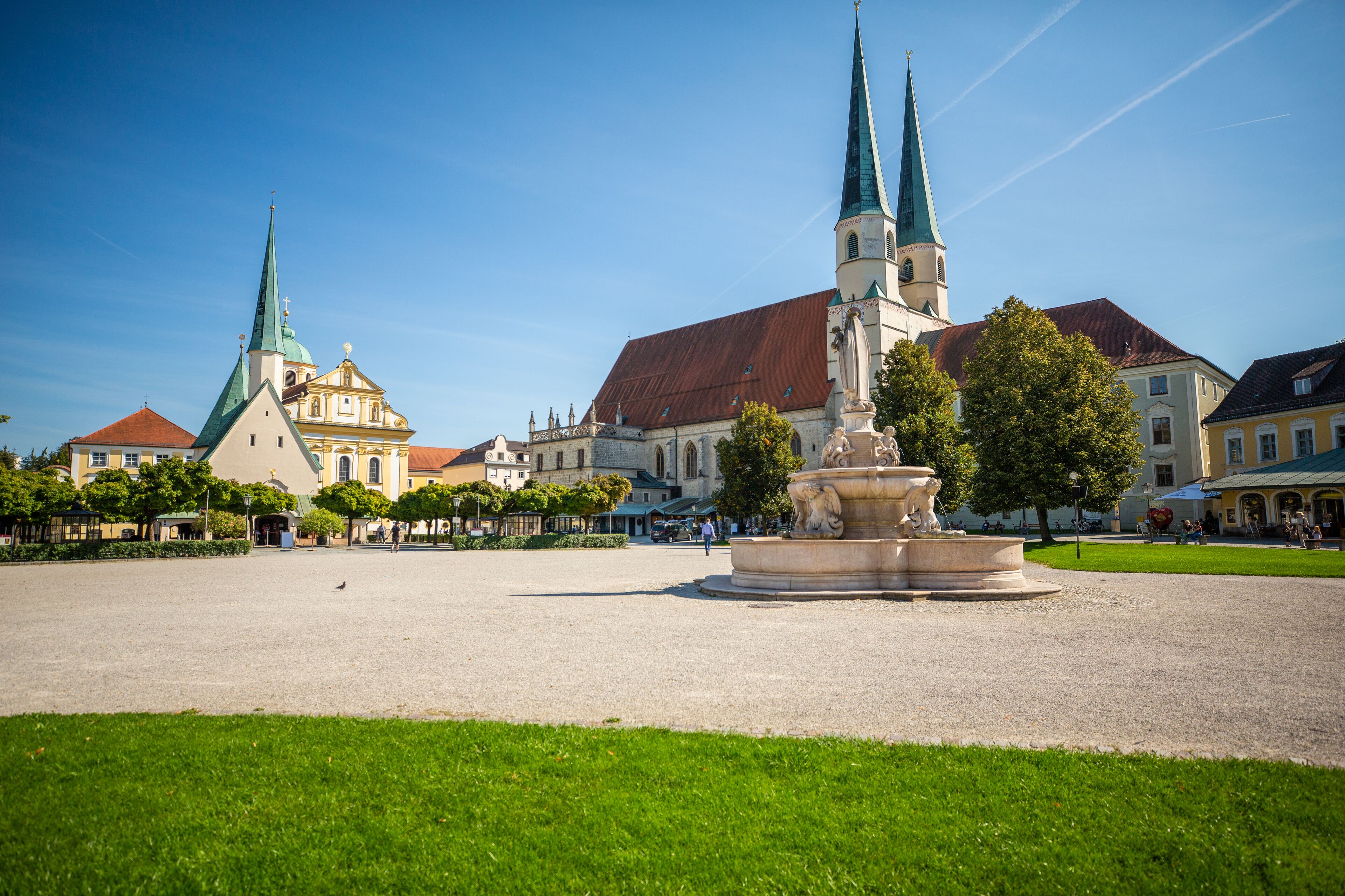 Ein Platz mit einer zentralen Fontäne, umgeben von historischen Gebäuden mit unterschiedlichen Baustilen, darunter eine Kirche mit einem grünen Kirchturm.
