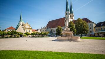 Ein Platz mit einer zentralen Fontäne, umgeben von historischen Gebäuden mit unterschiedlichen Baustilen, darunter eine Kirche mit einem grünen Kirchturm.