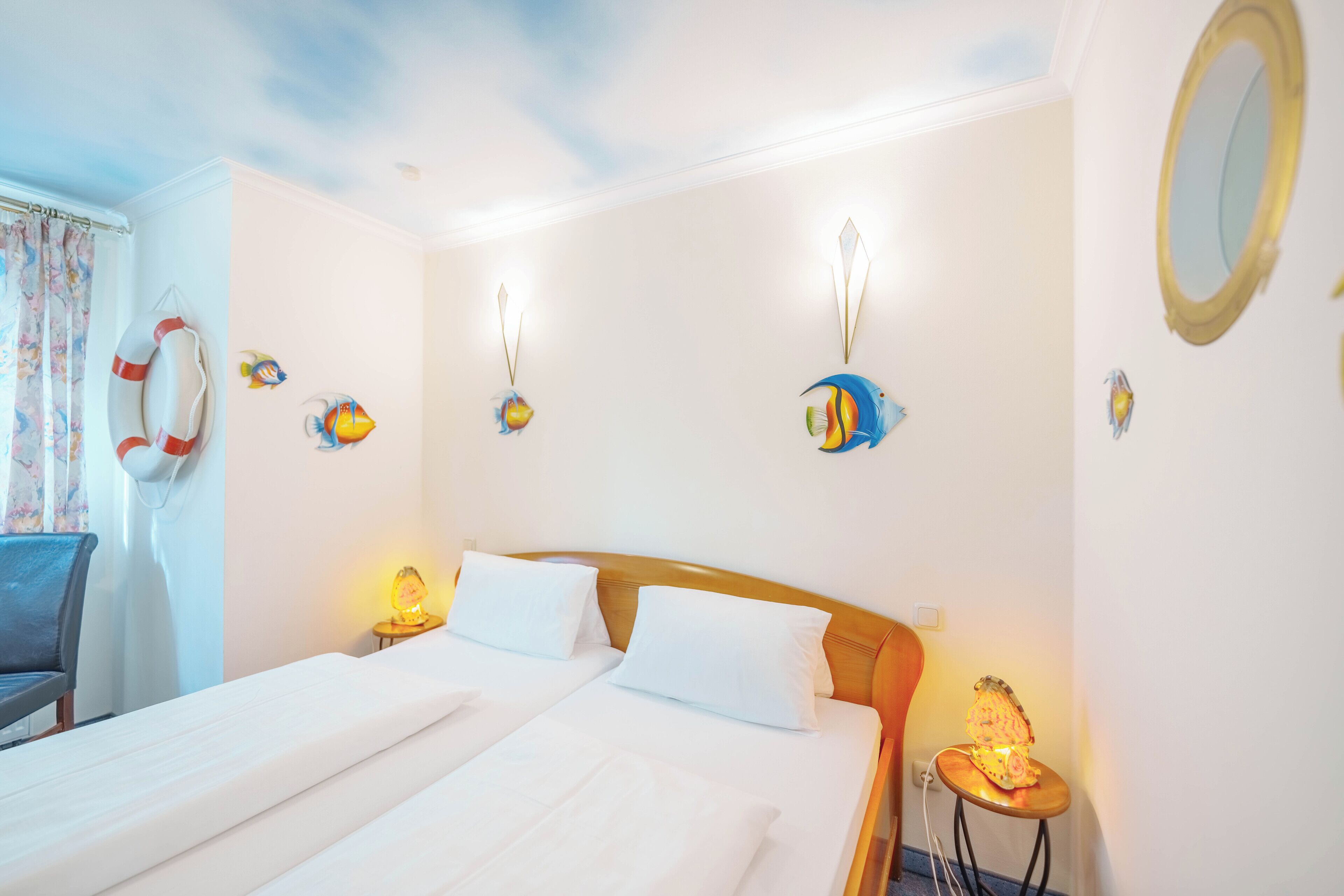 A hotel room with two beds, a wooden headboard, a small table with a lamp, and wall decorations including fish and a lifebuoy.