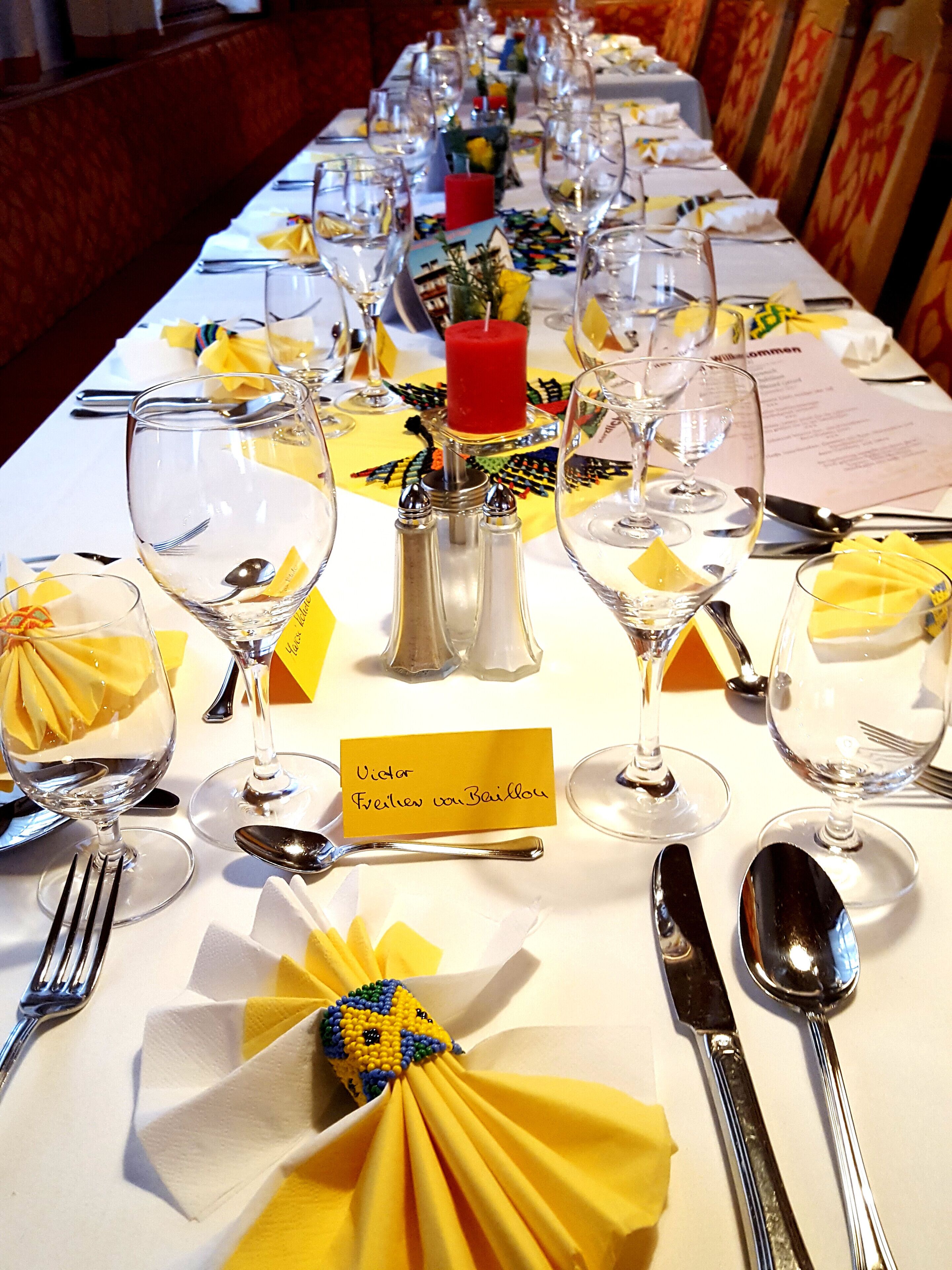 Una larga mesa de comedor dispuesta con múltiples copas de vino, tenedores y cucharas, cada una con un servilleta amarilla atada en lazo. También se observa una vela roja y un juego de salero y pimentero.