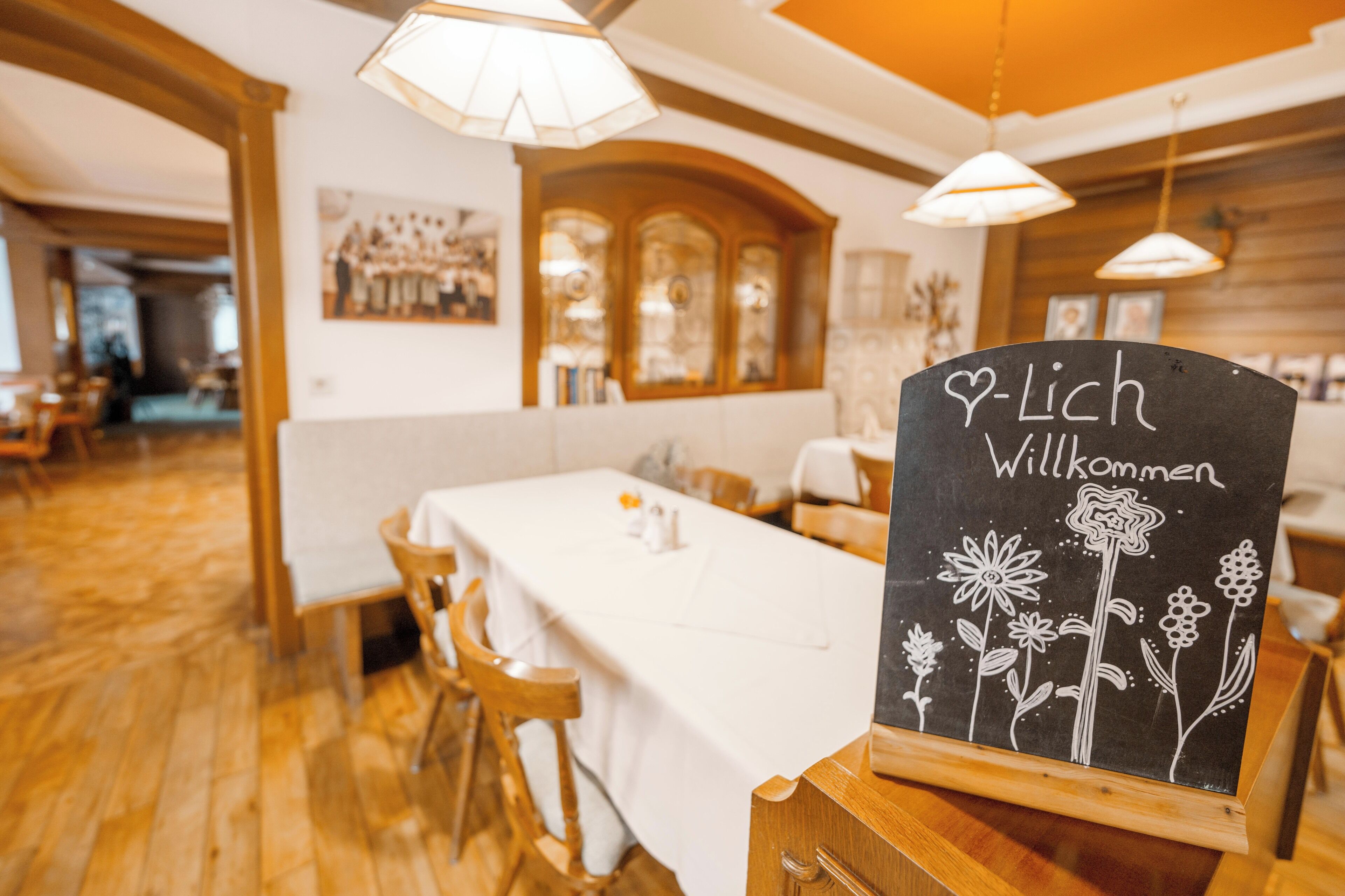 A restaurant interior with wooden floors, white tablecloths, and a chalkboard sign reading "Lich Willkommen".