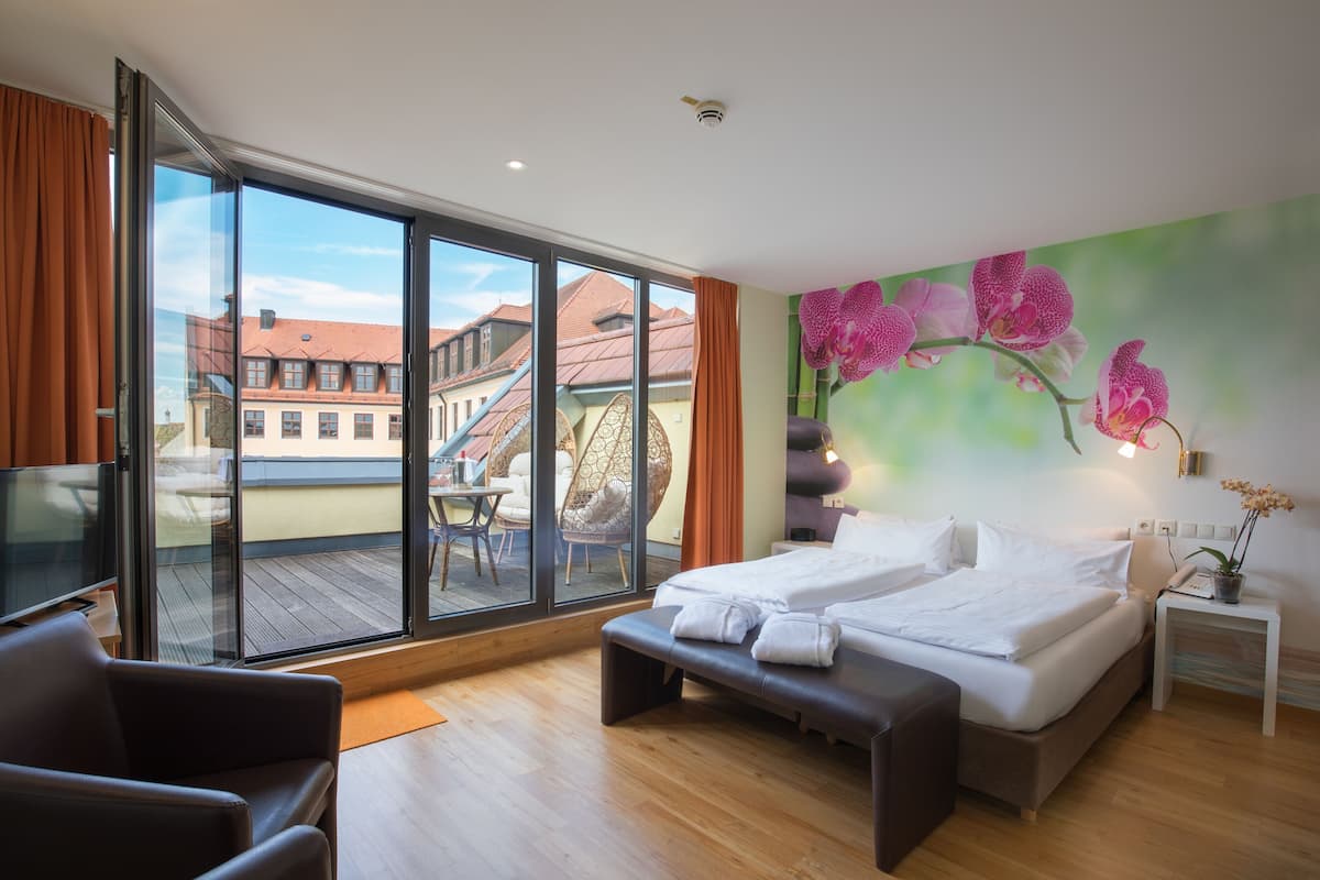 Une chambre d’hôtel dotée d’une grande porte-fenêtre coulissante donnant sur une terrasse extérieure, d’un lit avec deux oreillers, d’un banc, d’une petite table de chevet avec une lampe et d’un mur orné d’une fresque florale.