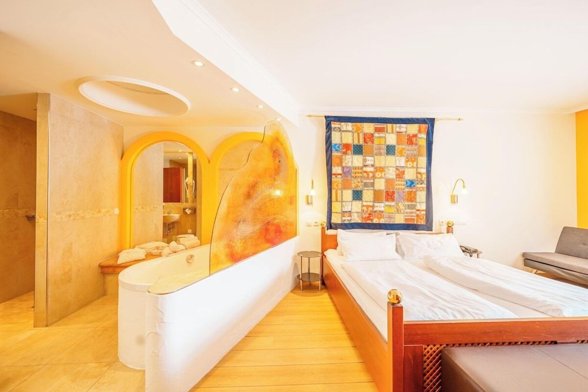 A hotel room with a wooden bed, a large mirror, a bathroom with a bathtub, and a quilted wall hanging.