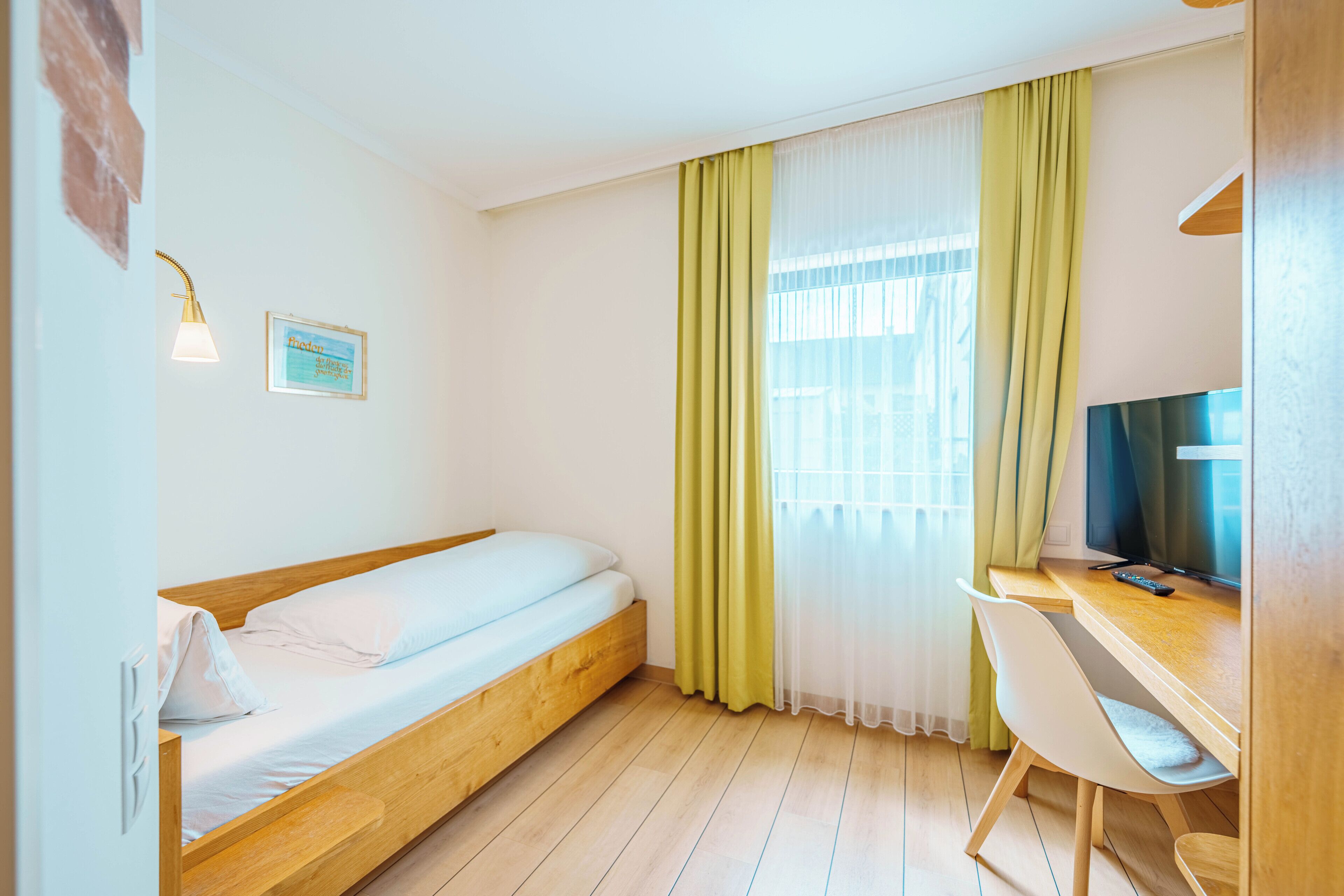 Quarto com cama de madeira, escrivaninha com cadeira, televisão e janela com cortinas.