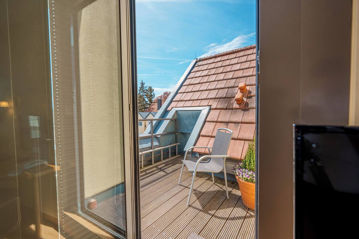 Un balcon avec une chaise et un pot de fleurs, une fenêtre avec des stores et une vue sur un immeuble doté d’un toit en pente.