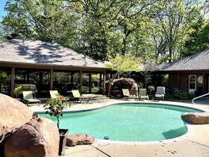 A covered outdoor pool area with lounge chairs, a brick building, and a large rock in the foreground. - The Courtyard Suite, Pool is opened in May and closed after Labor Day. (Joplin)