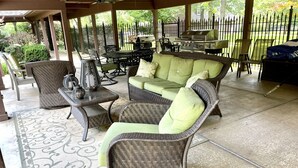Een overdekt terras met rieten meubilair, een barbecue en uitzicht op een omheinde tuin.