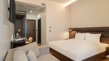 A modern hotel room with a large bed, a sofa, a desk with a lamp, and a mirror.