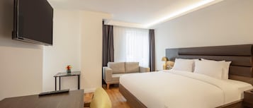 Una camera d'albergo con un letto, una TV fissata al muro, un tavolino con un telecomando, una poltrona e una finestra con tende.