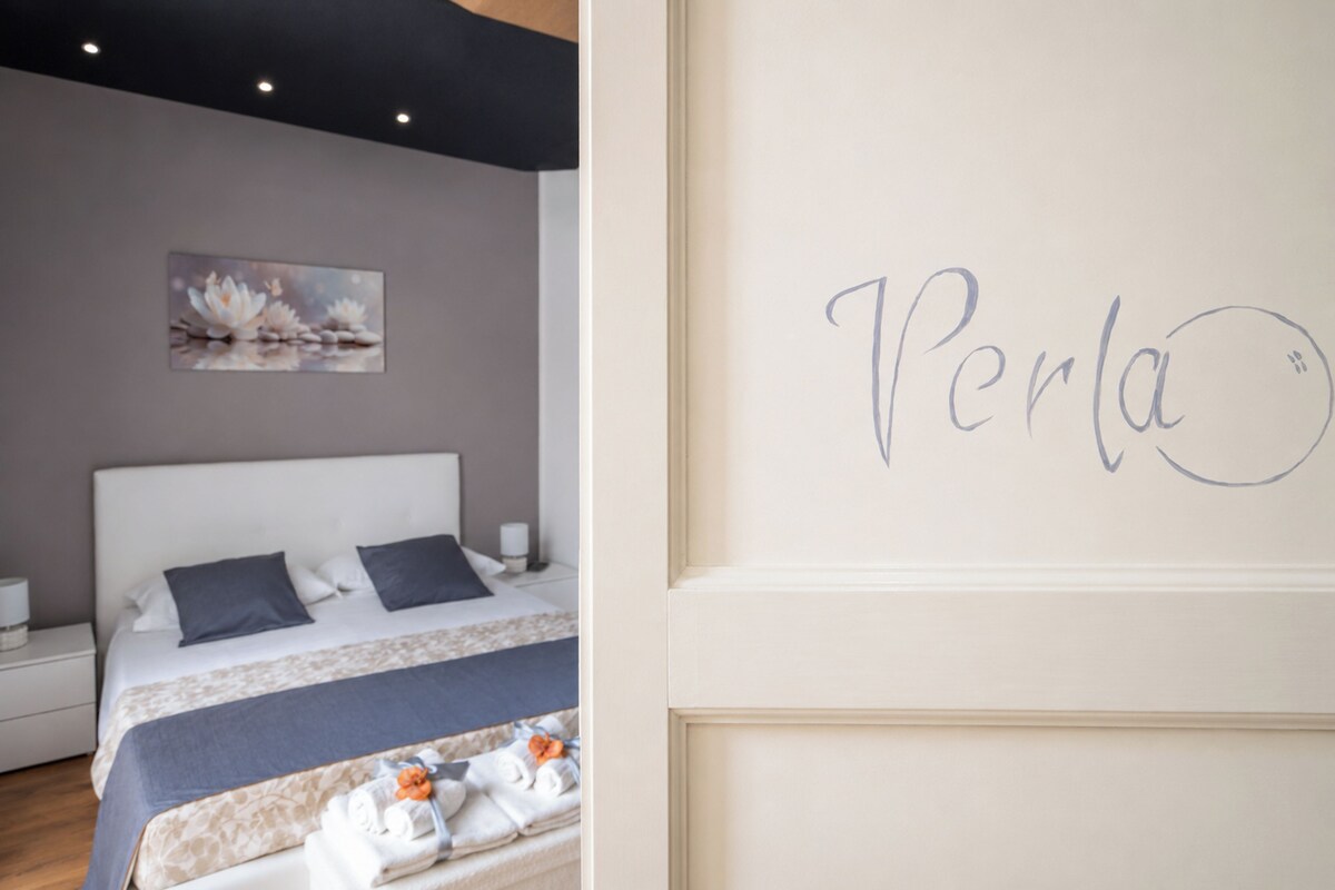 Quarto com cama, almofadas, mesa de cabeceira, abajur e parede com um nome.