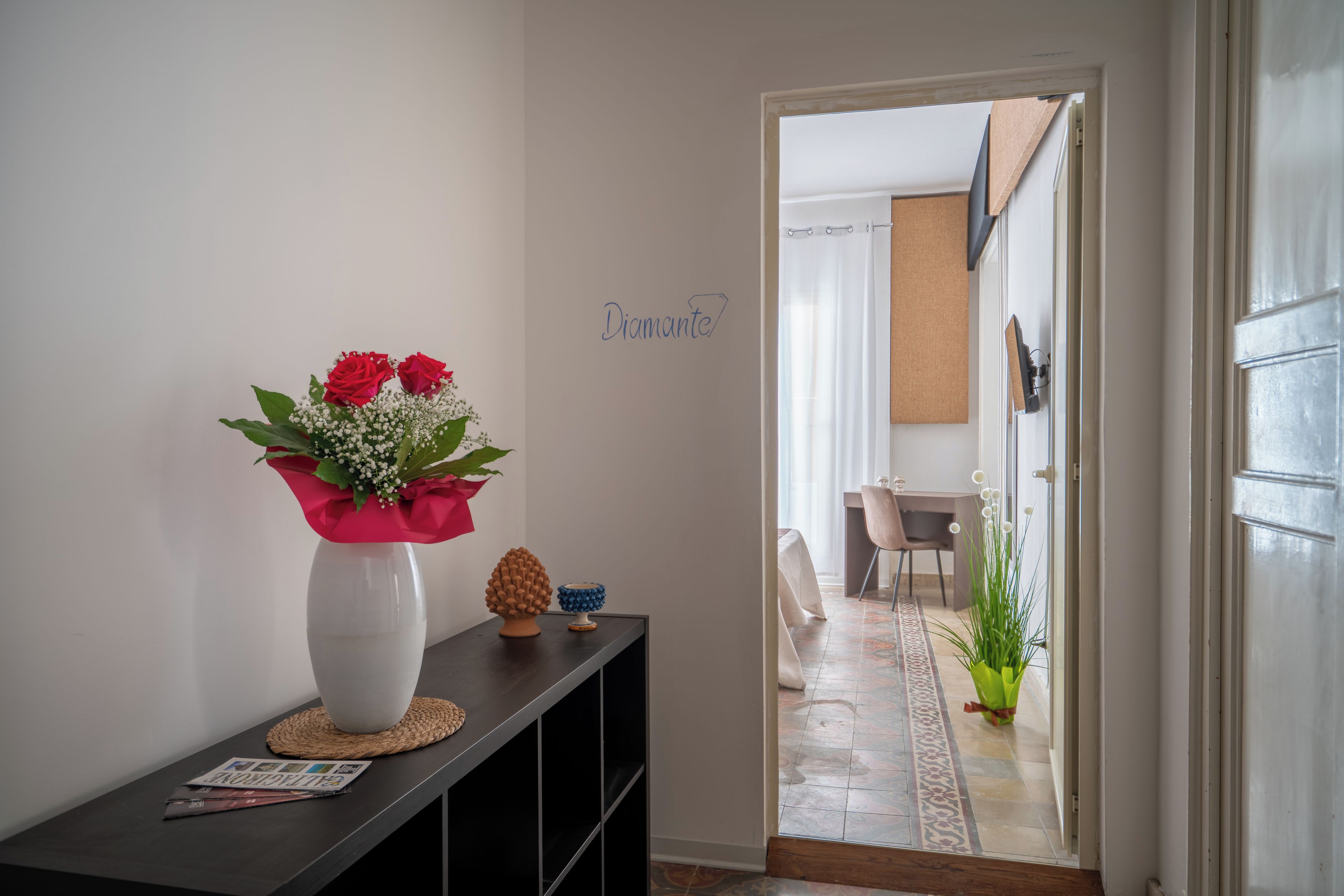 Um corredor com um vaso branco com flores vermelhas em uma estante, um pequeno pinhão e um objeto azul na estante, e uma porta com uma placa que diz 'Diamond'.