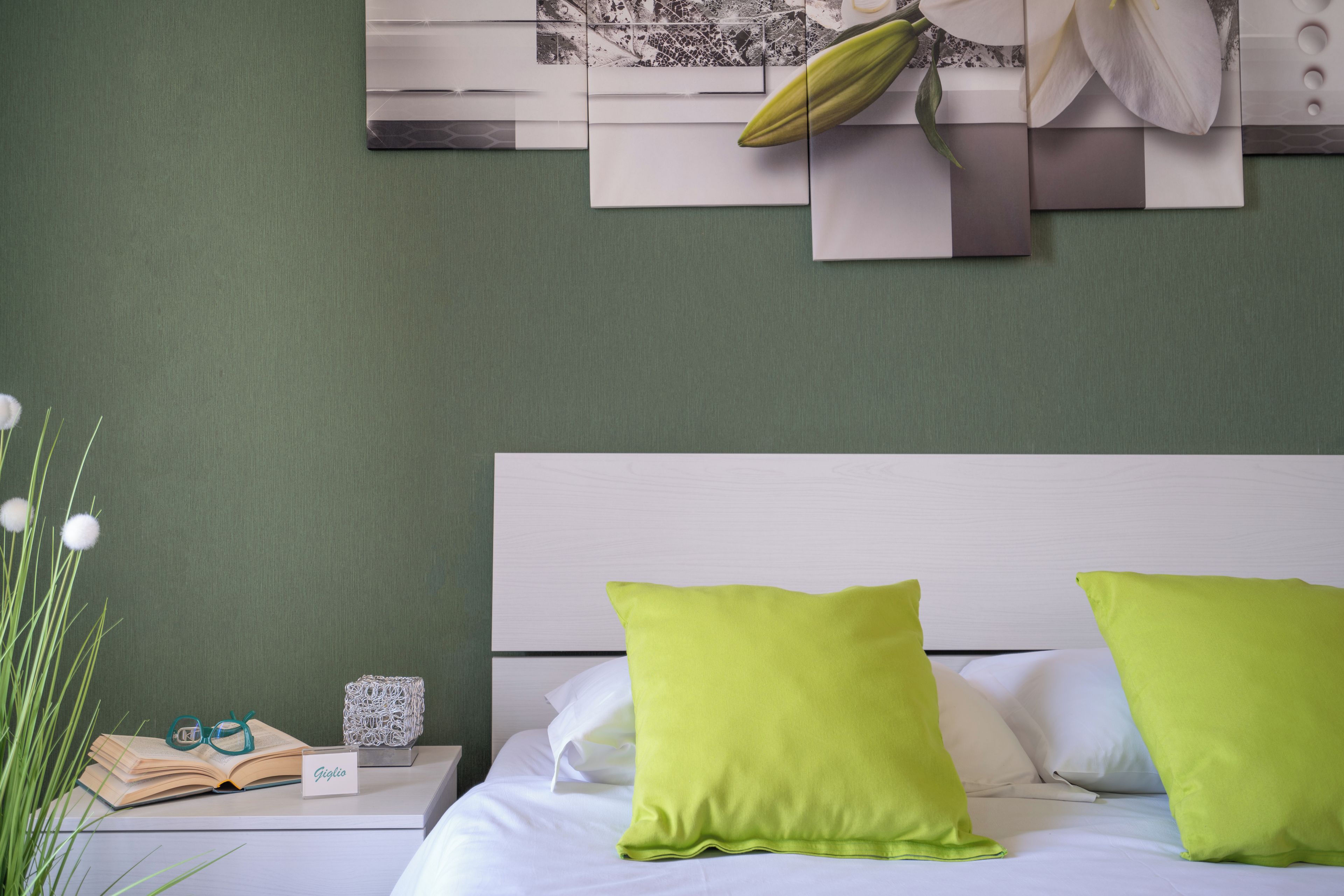 A neatly made bed with two pillows, a bedside table with a book and glasses, and a wall with a framed picture and a plant.