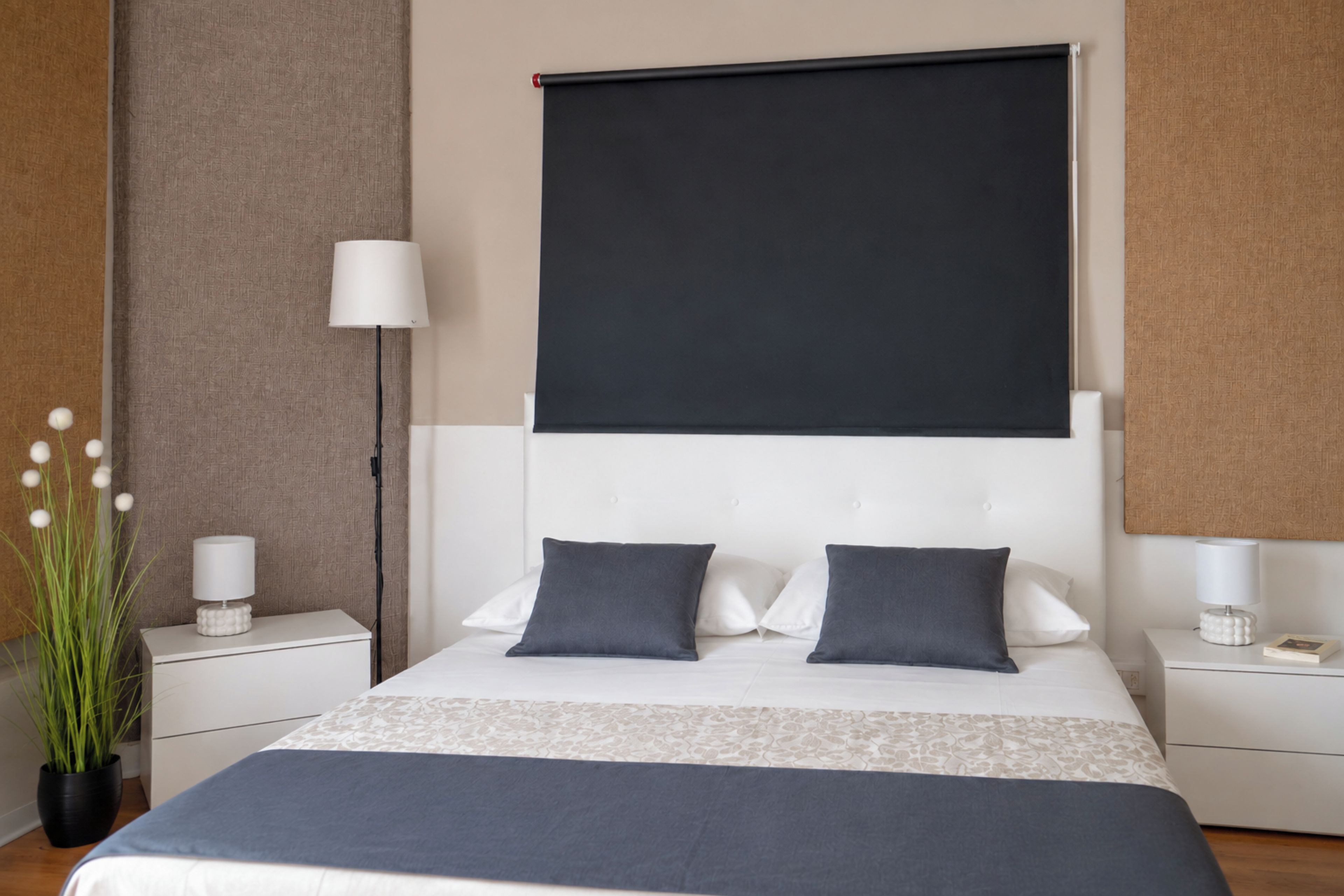 A modern hotel room with a large bed, two bedside tables, a nightstand, a wall-mounted lamp, and a potted plant.