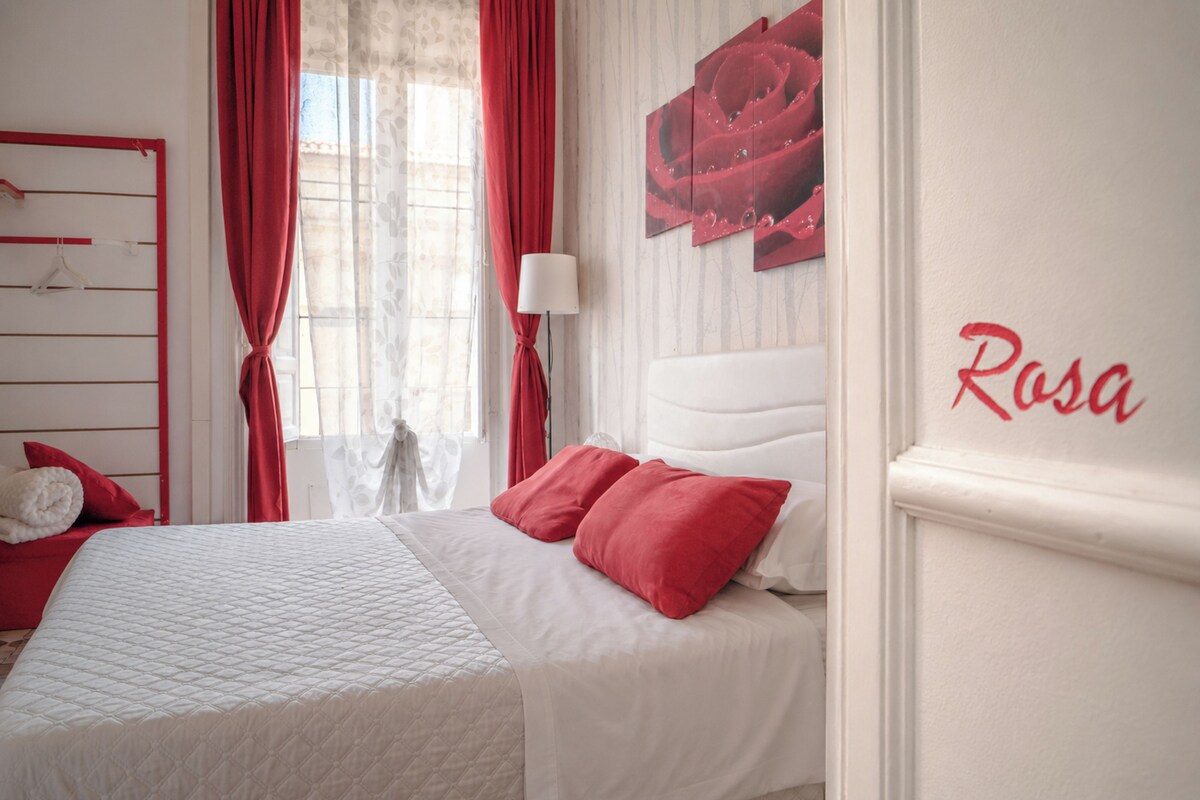 Quarto com cama, cortinas vermelhas, parede branca com um nome e janela com cortinas finas.