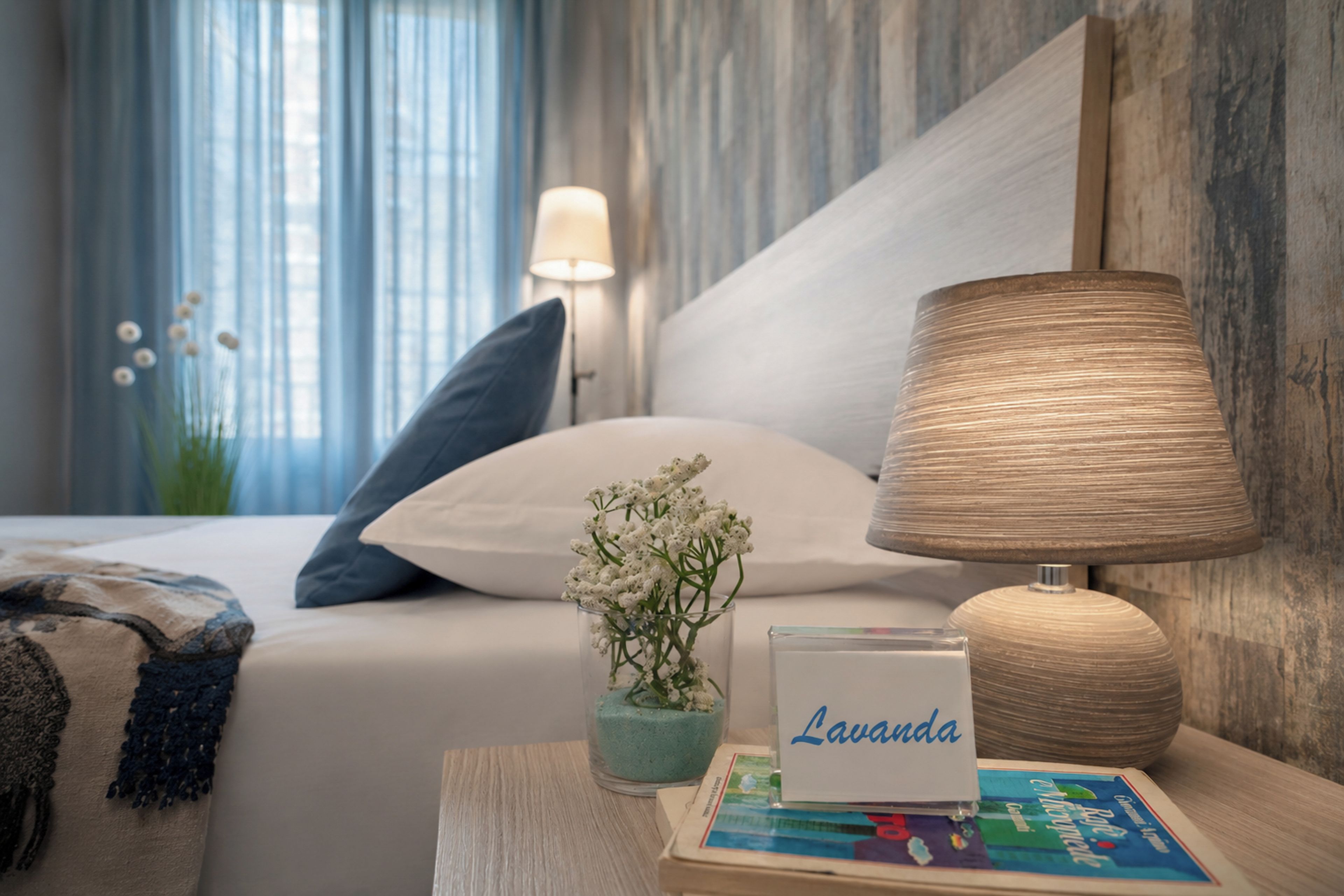 Ein ordentlich bezogenes Bett mit weißer Bettwäsche und einem blauen Kopfkissen, eine Nachttischlampe, eine kleine Vase mit weißen Blumen und ein Schild mit dem Wort ‚Lavanda‘.