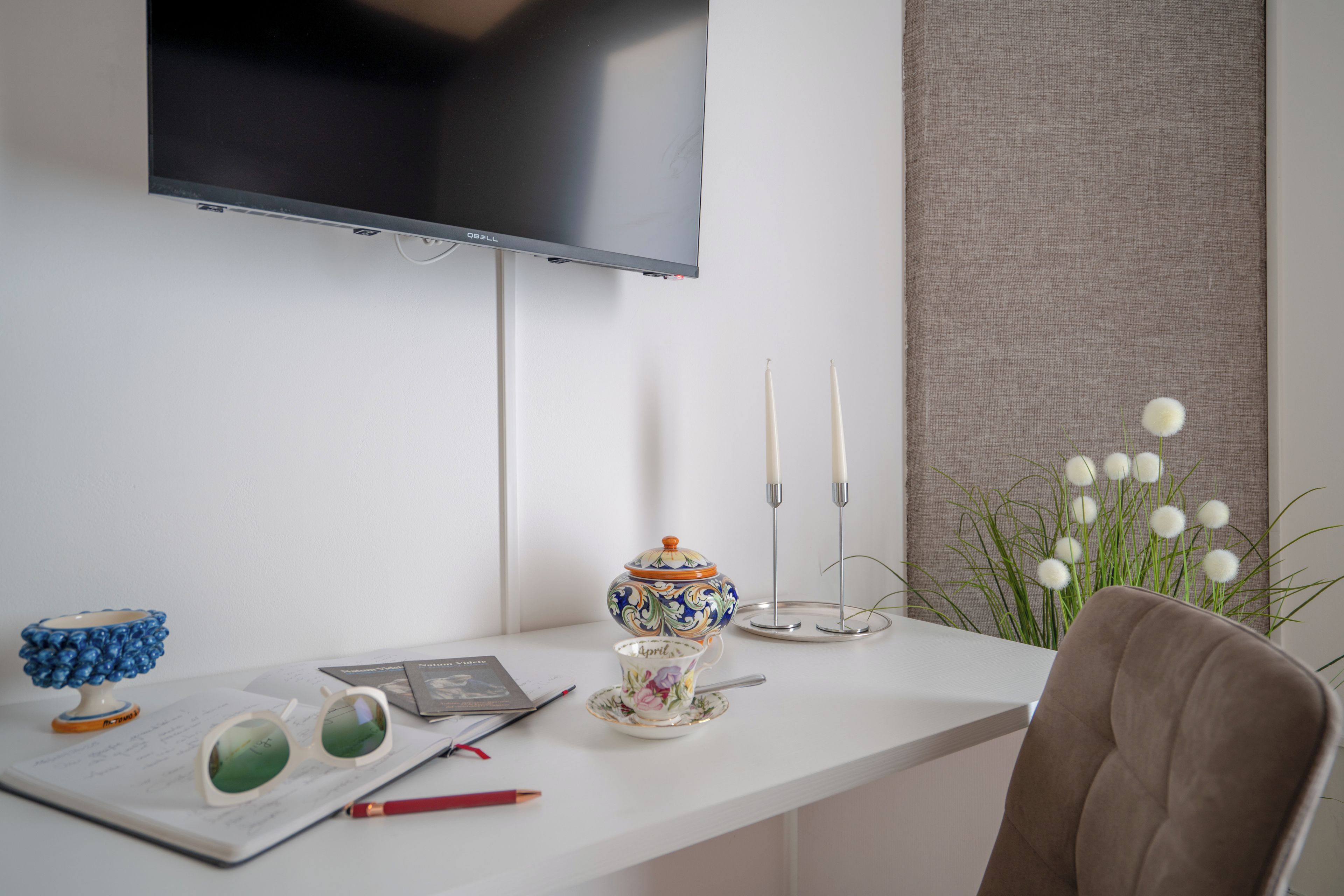 Ein ordentlich eingerichteter Schreibtisch mit einem darüber montierten Fernseher, einer Vase mit weißen Blumen und einem Stuhl.