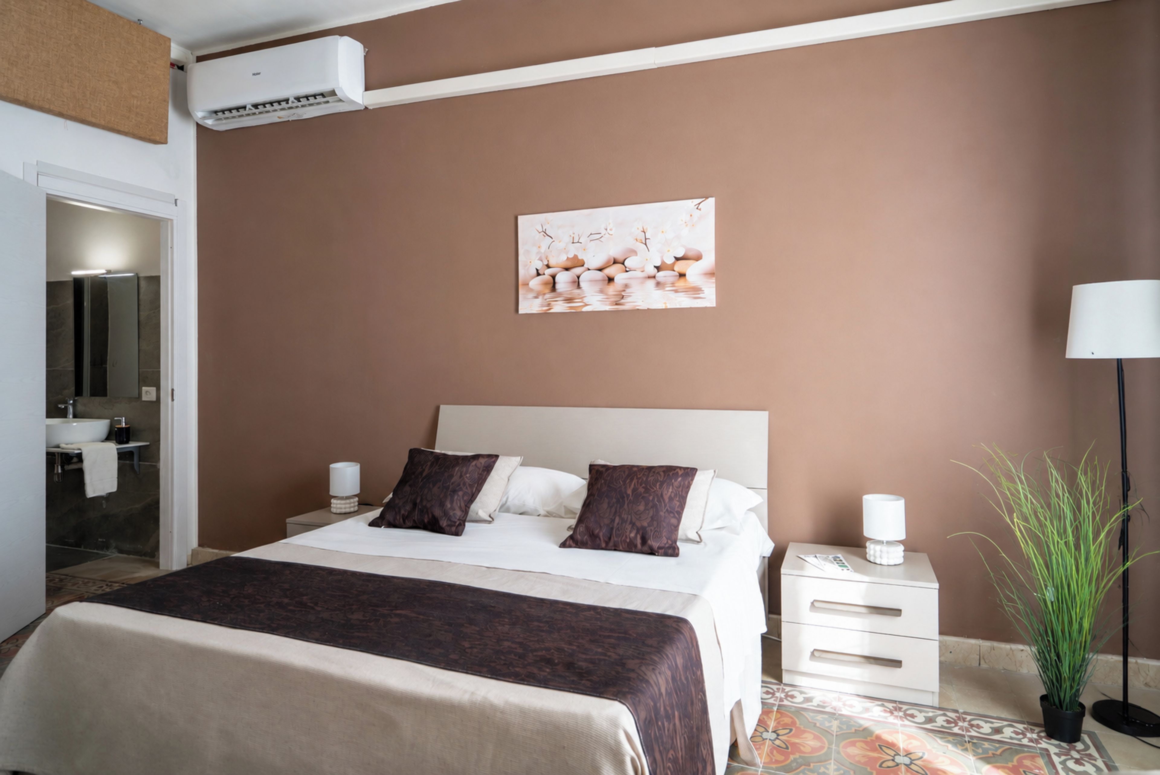 A hotel room with a bed, bedside tables, a nightstand, a potted plant, and a wall-mounted air conditioner.