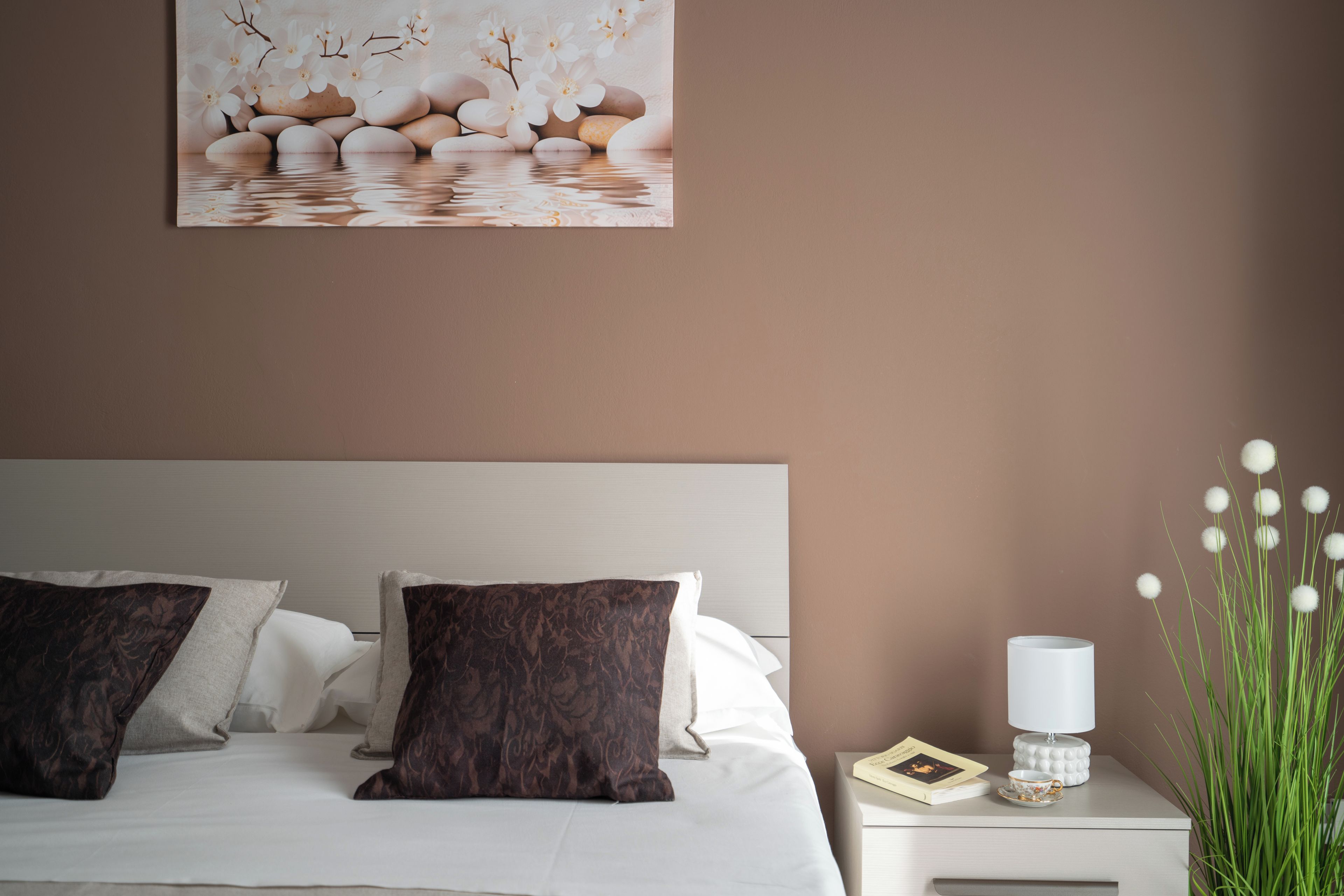 Une chambre à coucher avec un lit, des oreillers, une table de chevet avec une lampe, un livre et un vase avec des fleurs.