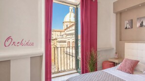 A hotel room with a bed, a small bedside table, a nightstand, a potted plant, and a view of a building with a dome through a window with pink curtains. - I Colori della Sicilia (Caltagirone)
