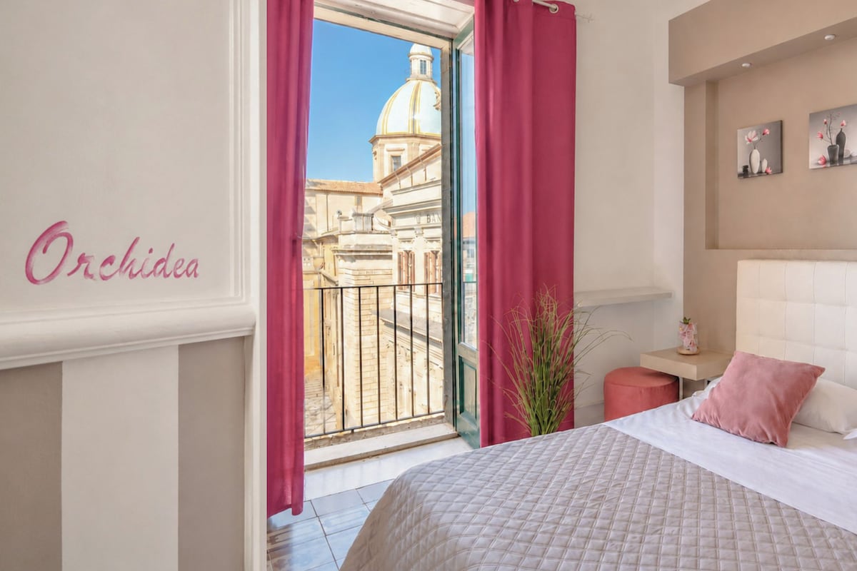 Quarto de hotel com cama, mesa de cabeceira, planta em vaso e vista para um edifício com cúpula através de uma janela com cortinas cor-de-rosa.