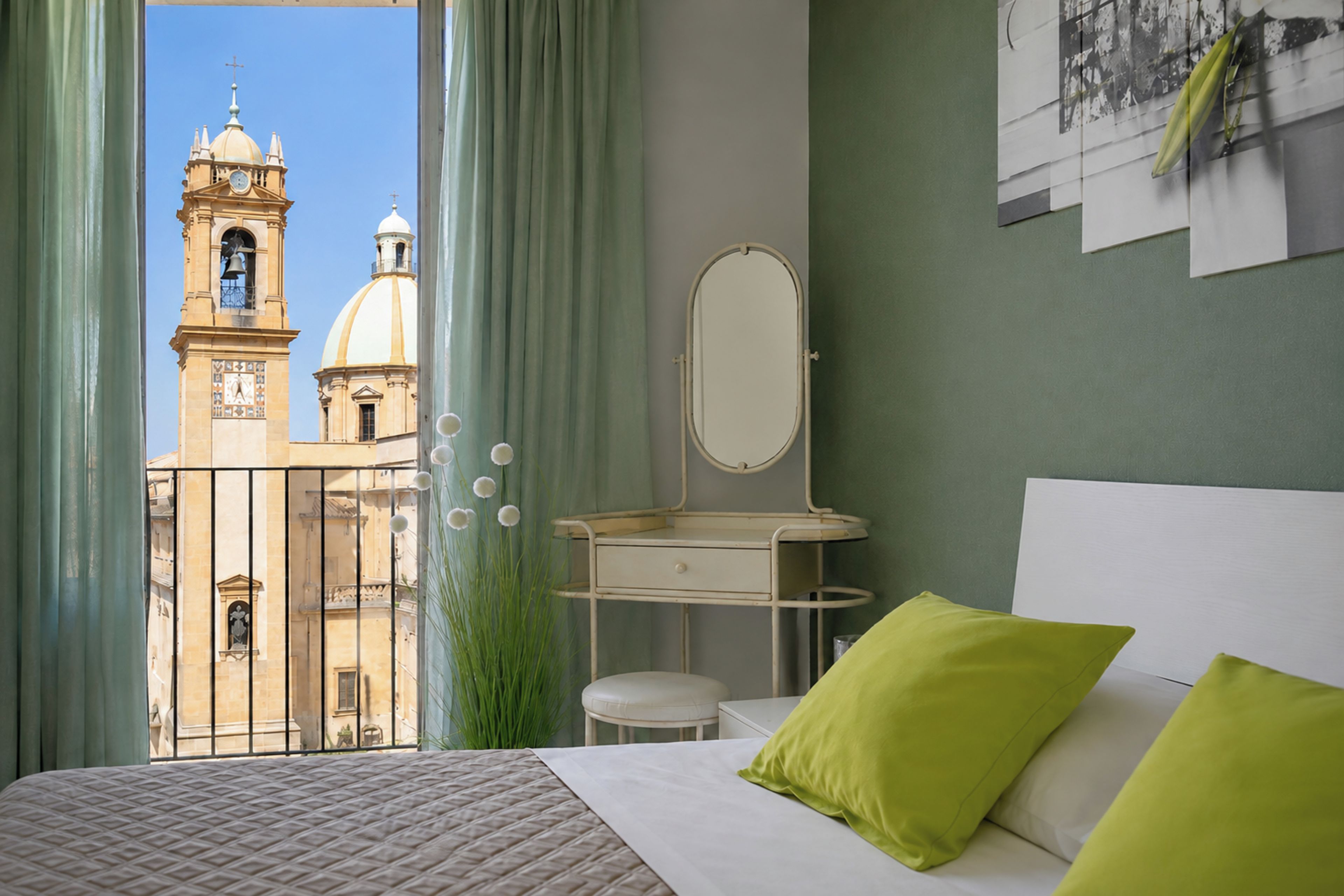 A hotel room with a bed, a bedside table with a mirror, and a view of a building with domes and a bell tower.