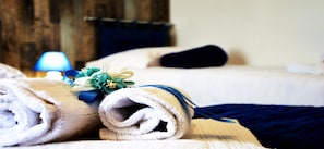 Uma cama bem arrumada com uma toalha branca enrolada e uma fita azul, um cobertor azul escuro e uma pequena planta decorativa com flores amarelas.