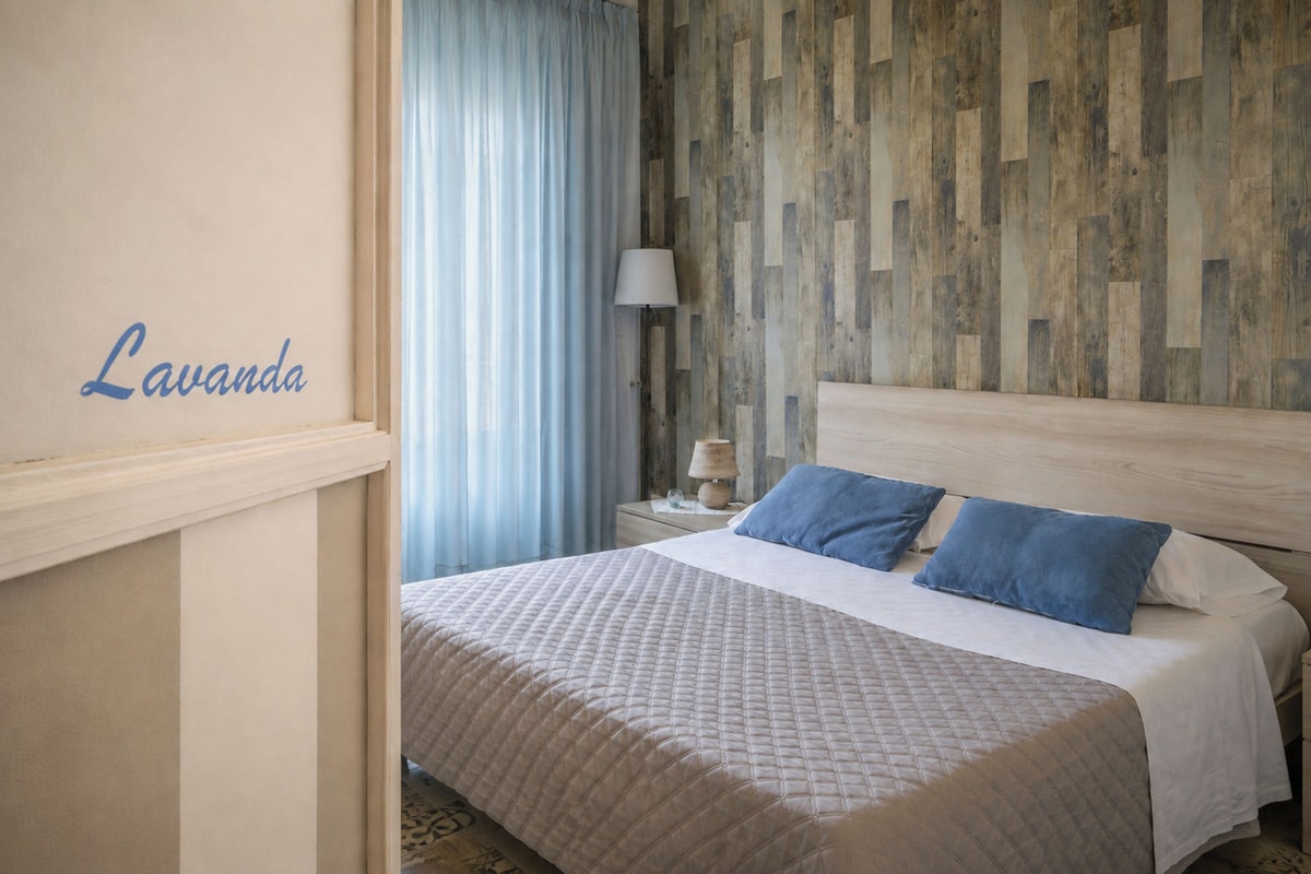 Quarto de hotel com cama, mesa de cabeceira, luminária e um painel na parede.