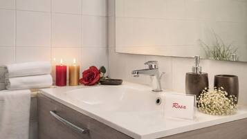 Un baño con un lavabo blanco, un espejo, un perchero para toallas, una vela roja, una vela dorada, una rosa roja y un letrero con la palabra 'Rosa'.