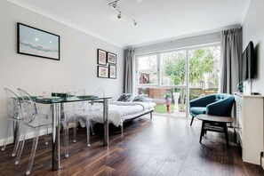 Uma sala de estar moderna com uma mesa de jantar de vidro, cadeiras transparentes, uma cama, um poltrona azul e uma TV fixada na parede.