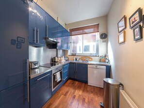 A compact kitchen with blue cabinets, a stainless steel range, a dishwasher, and a window with blinds. - Inviting 2BD Victorian Flat, Near Hampstead (London)