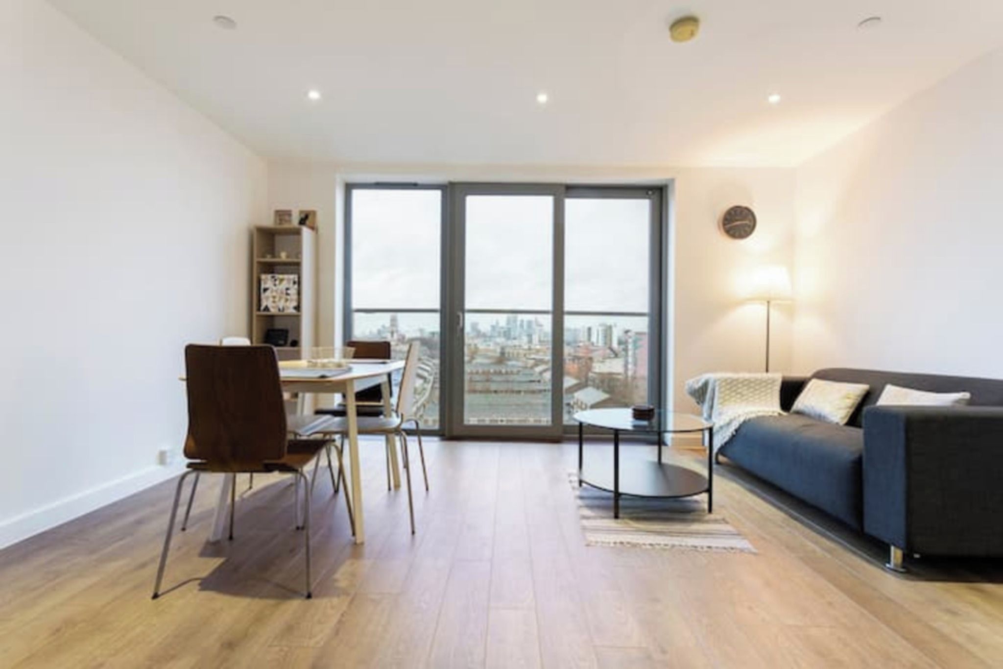 A modern living room with a sofa, a dining table with chairs, and a view through a large window.