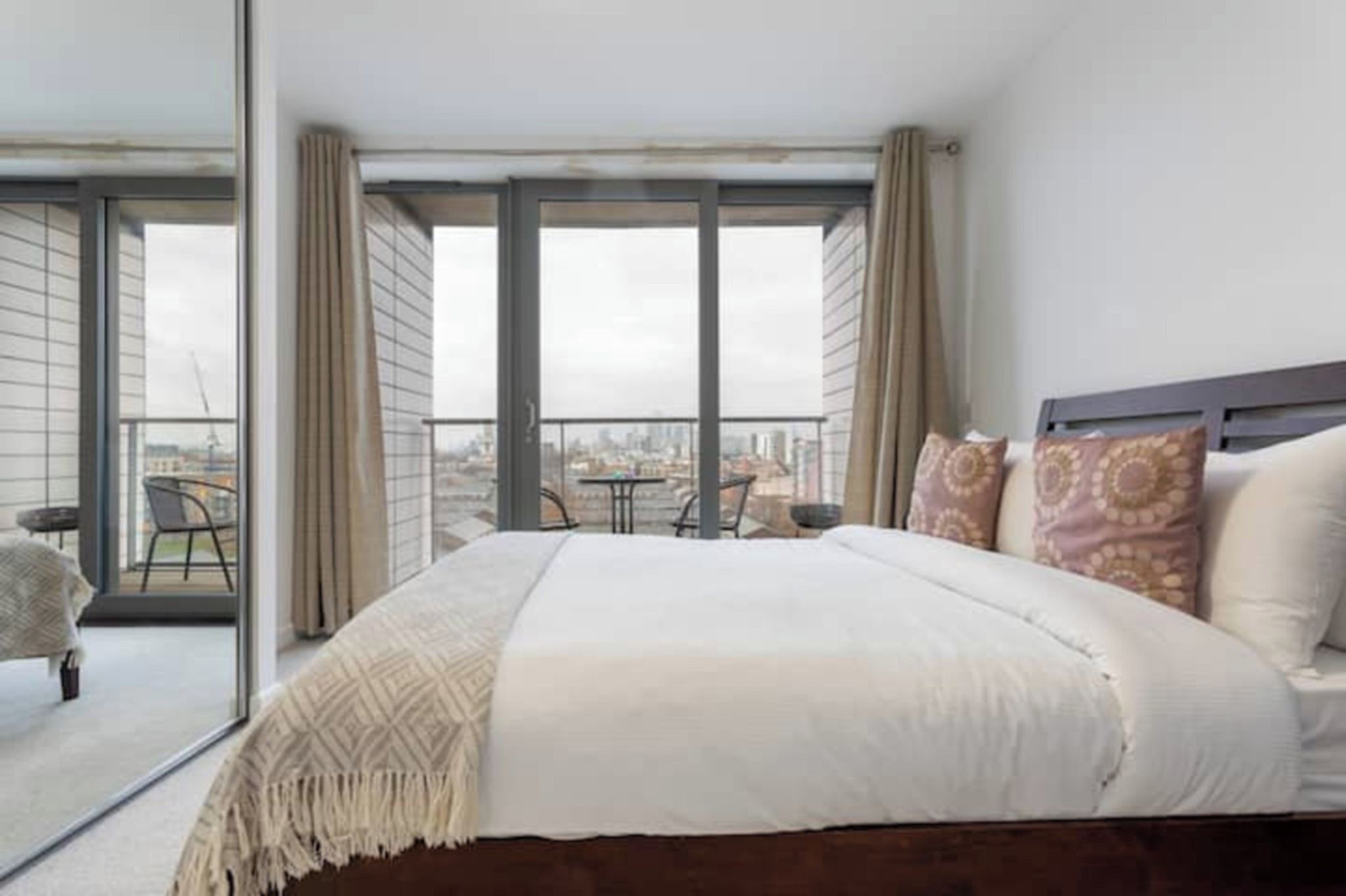 Un dormitorio con una cama grande, un balcón con mesa y sillas, y vista a la ciudad.