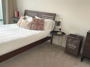 Un lit bien fait, avec des oreillers, une table de chevet avec une lampe, et un sac de voyage étiqueté « linge sale » posé sur le sol.