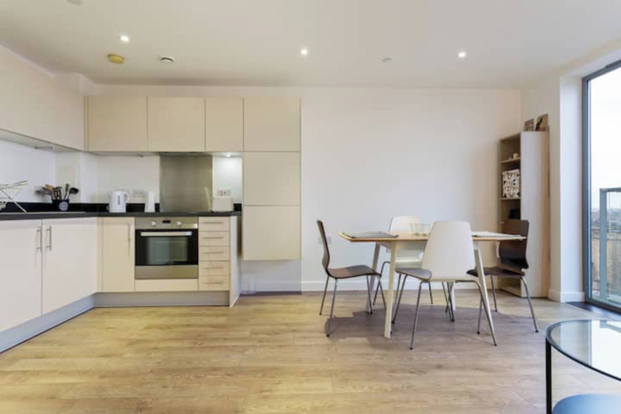 A modern kitchen with a dining area, wooden flooring, and a view through a large window.