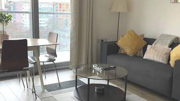 A modern living room with a glass coffee table, a sofa with cushions, a dining area with a table and chairs, and a large window offering a city view.